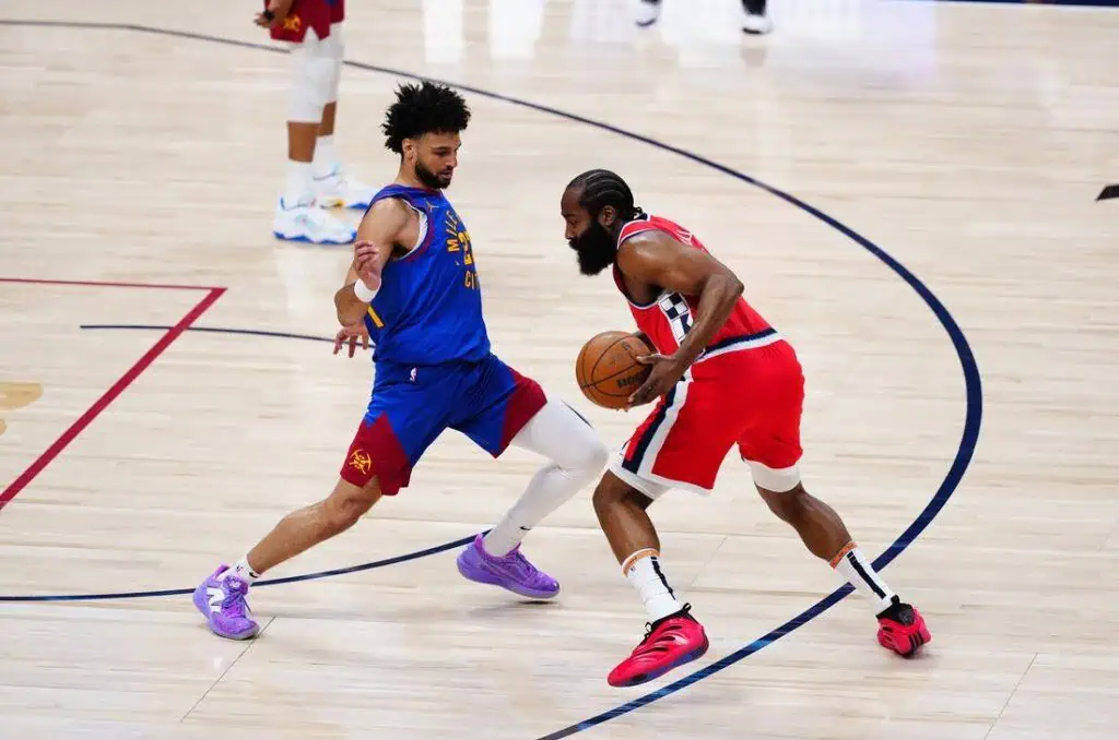 A basketball player in a red and white uniform dribbles while being guarded by an opponent in a blue jersey during a dynamic game scene.