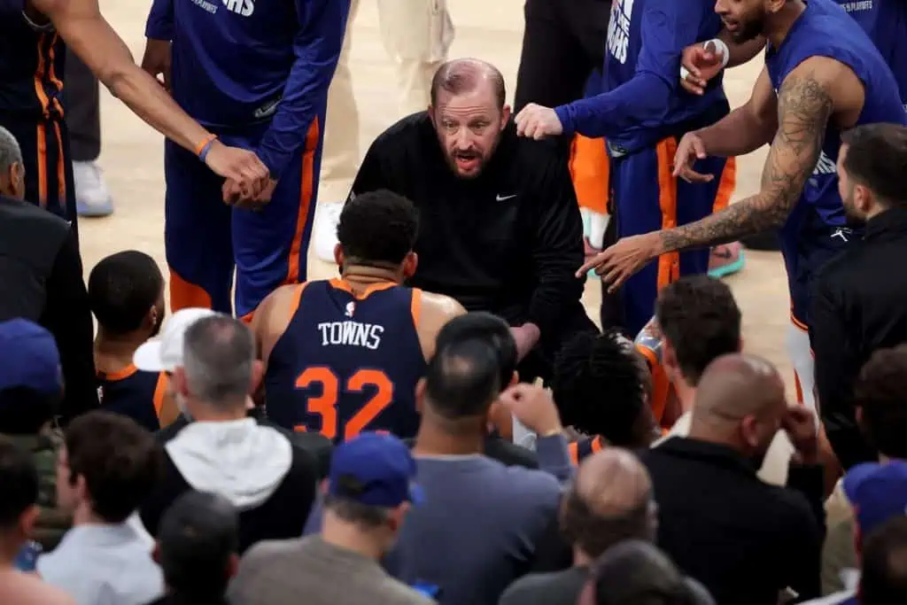 A huddle of basketball players surrounds a seated individual, with fans intently watching the scene unfold in an arena.