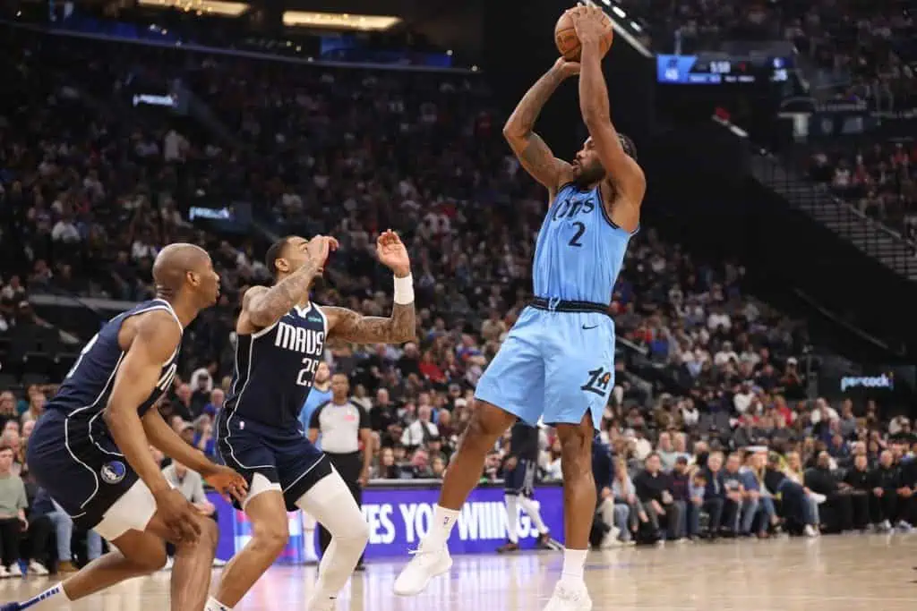6 A basketball player in a light blue uniform takes a jump shot, while two defenders in dark uniforms attempt to block him.