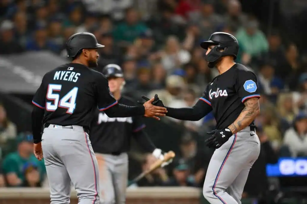 Two baseball players in black uniforms share a handshake and celebrate a play on the field, with fans visible in the background.