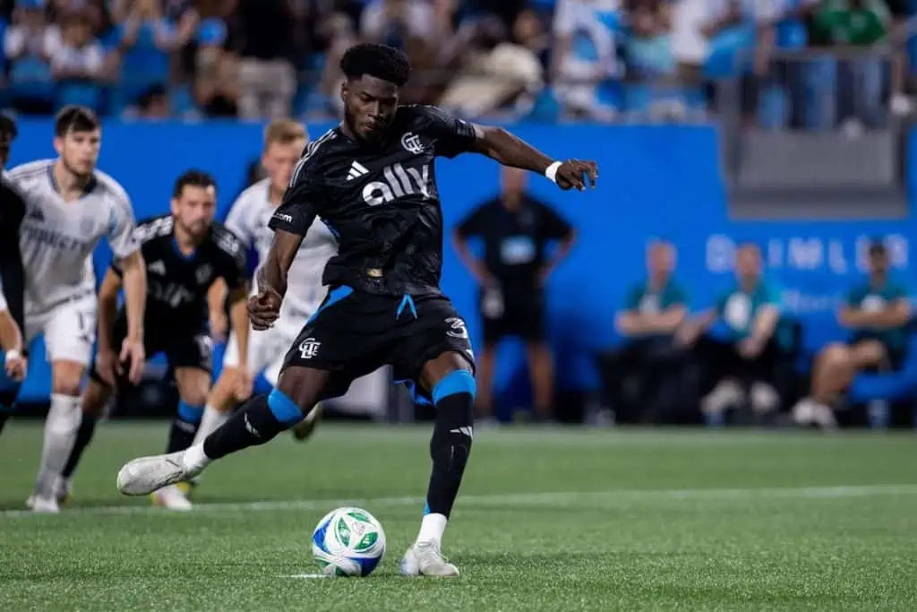 A soccer player in a black kit prepares to take a penalty kick, with teammates and opponents watching intently in a stadium setting.
