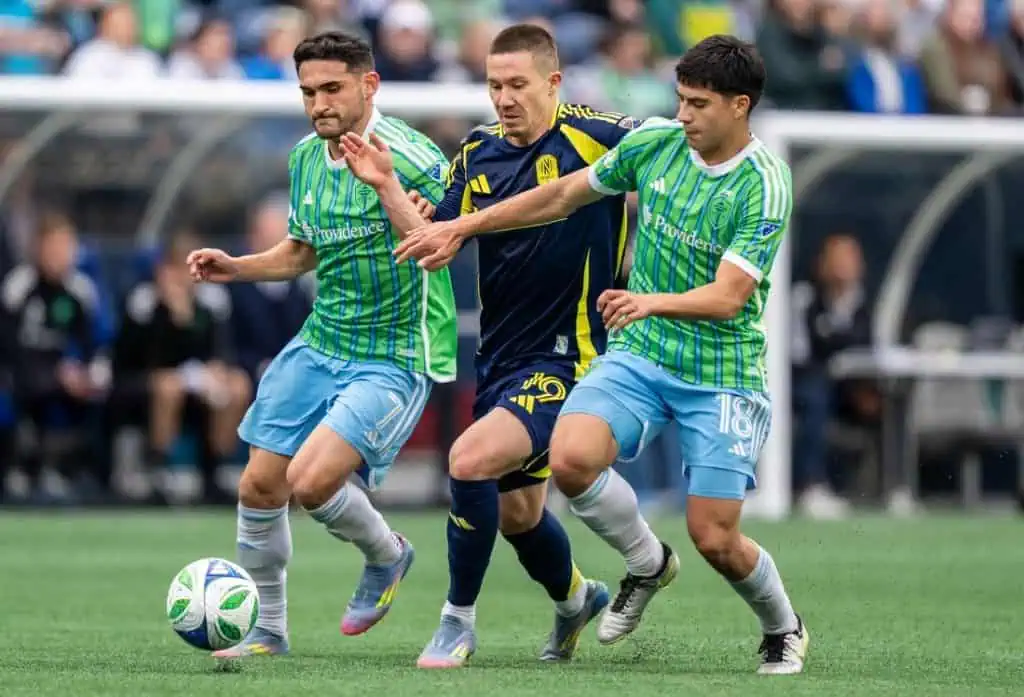 Two soccer players in green and blue jerseys are engaged in a competitive match, attempting to gain control of the ball from an opponent.