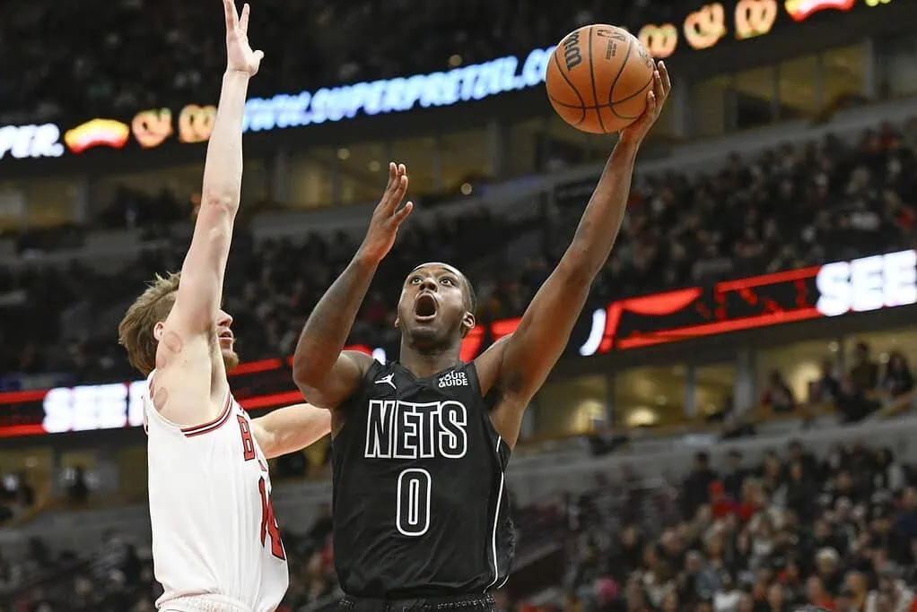 5 A basketball player in a black Nets jersey shoots a jump shot, while an opponent reaches to block the attempt in a packed arena.