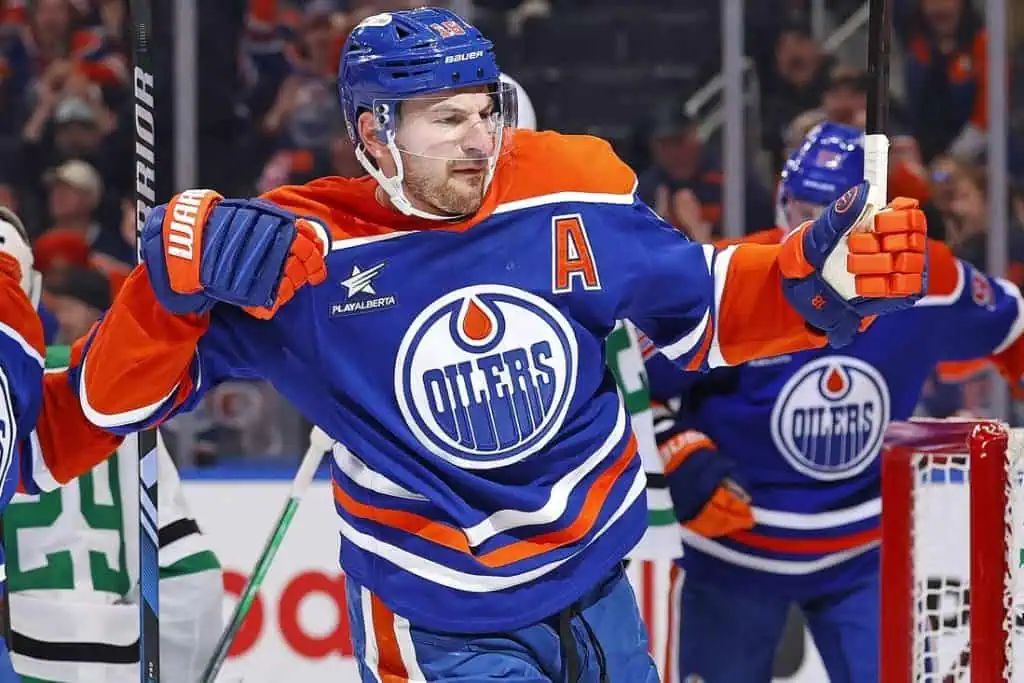 A hockey player in a blue and orange Edmonton Oilers jersey celebrates a goal, raising his arms in excitement on the ice.