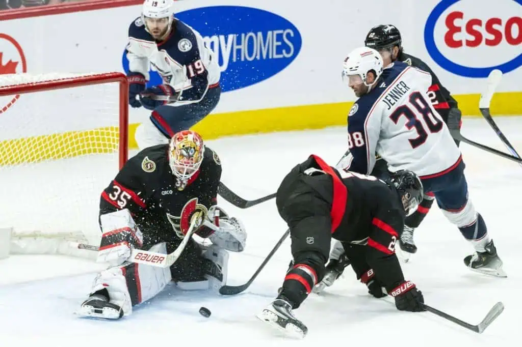 A tense moment in a hockey game with players battling for the puck near the goal, while the goalie prepares for a shot.