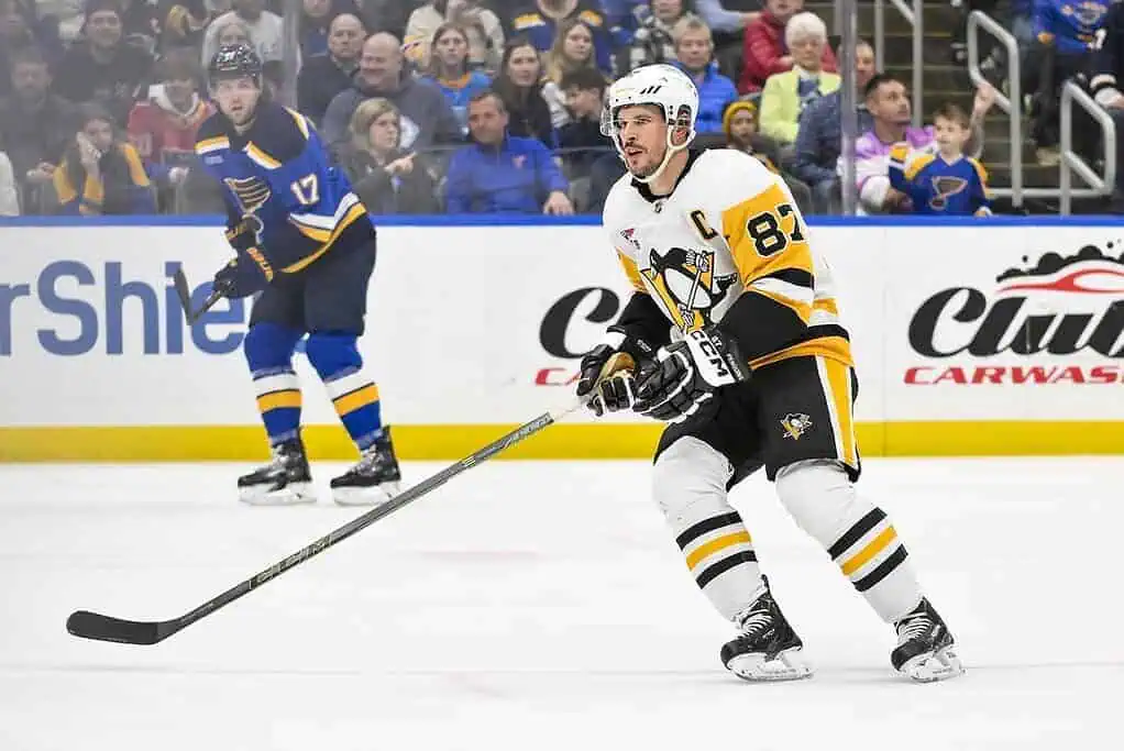 A hockey player in a black and gold uniform looks ready to pass the puck while another player, wearing blue, skates in the background.