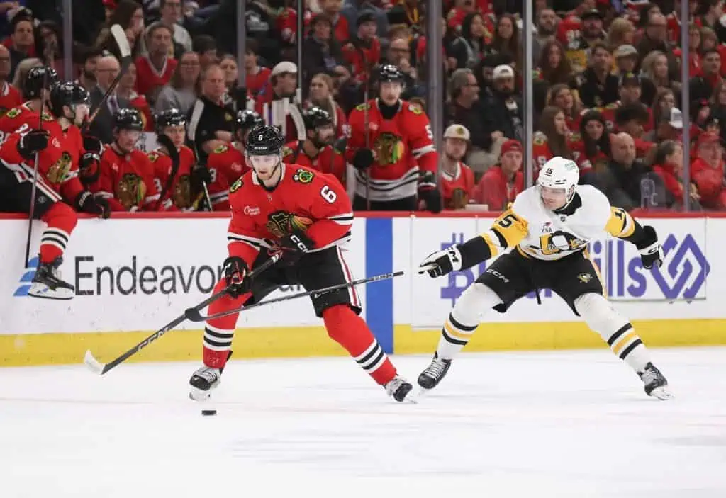 A hockey player in a red jersey dribbles the puck past an opponent in white while a cheering crowd in red jerseys watches.