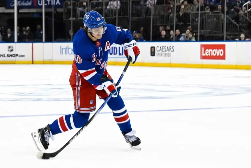 A hockey player in a blue and red uniform skating on the ice, preparing to take a shot with a focused expression.