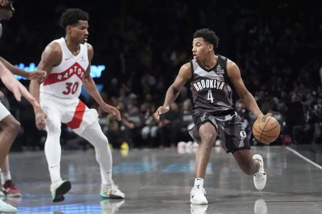 2 A player in a black Brooklyn jersey dribbles a basketball, while two defenders in white Raptors jerseys closely guard him on the court.
