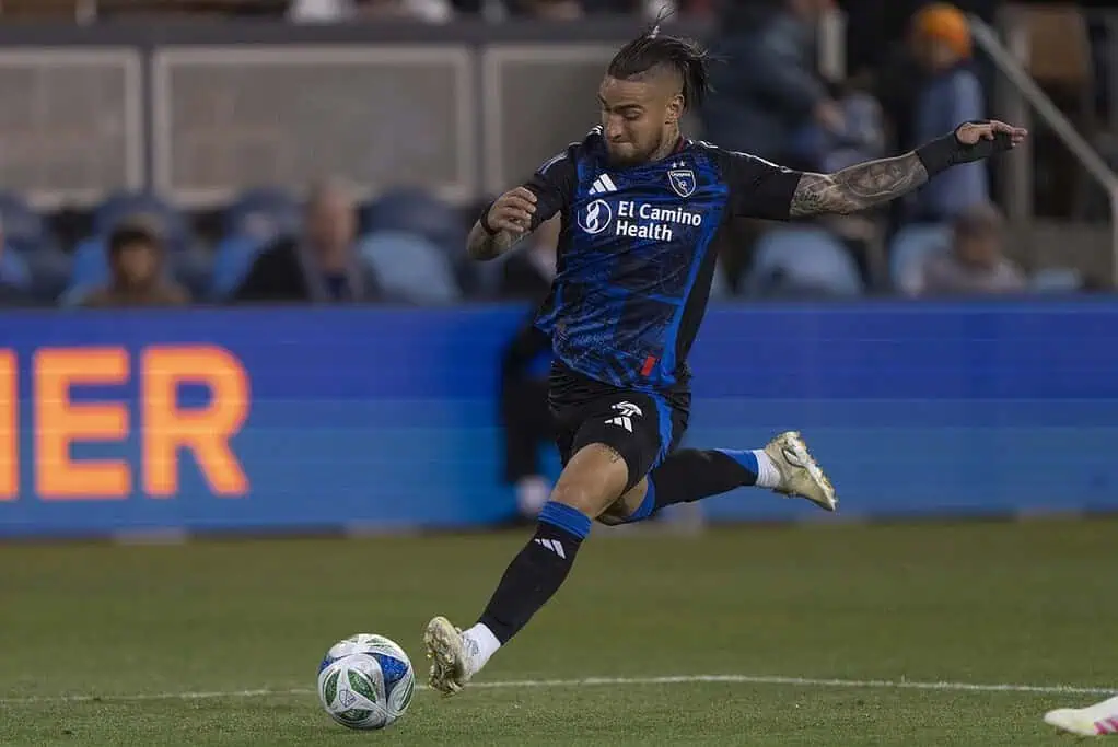 A soccer player in a blue and black uniform kicks a ball on the field during a match, with fans visible in the background.
