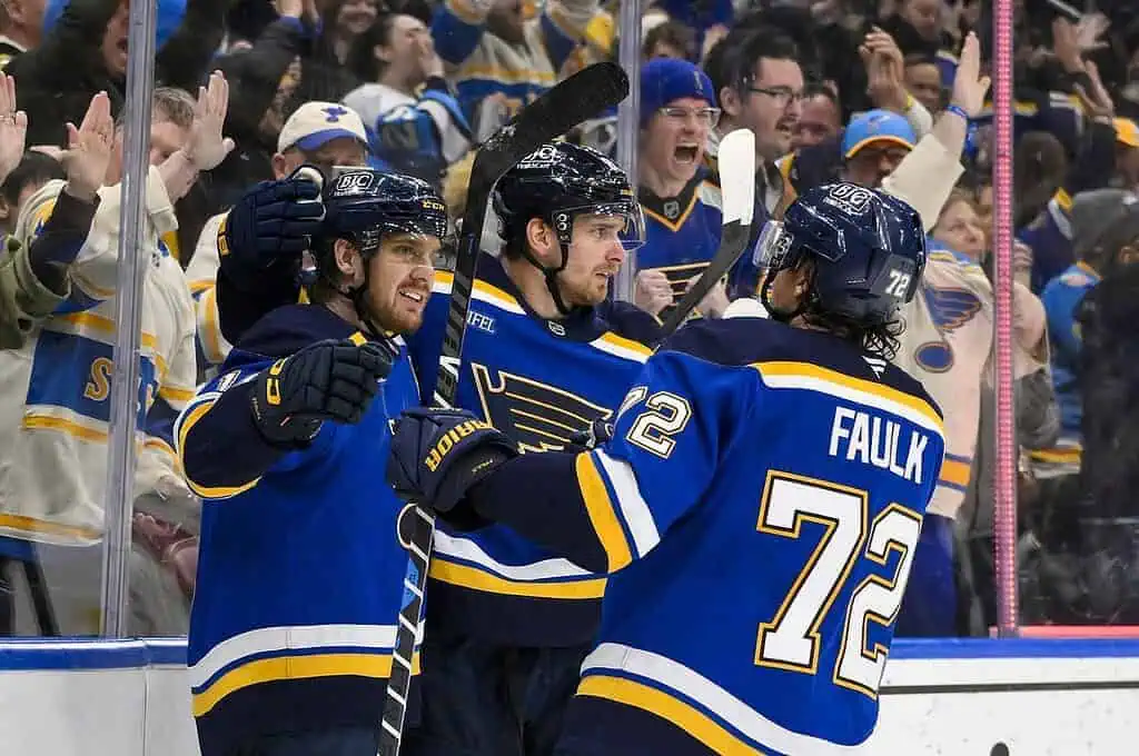 Three hockey players in blue jerseys celebrate enthusiastically, while fans in the background cheer during a game.