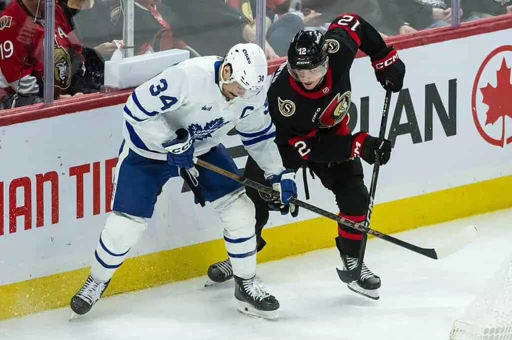 NHL 4 Two hockey players battle for the puck along the boards during a game, with one wearing a blue jersey and the other in black.