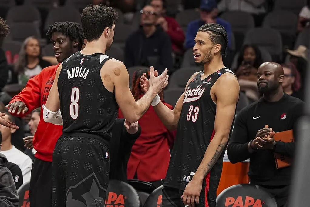 2 Two basketball players in black jerseys shake hands on the court, surrounded by teammates and fans in the stands.