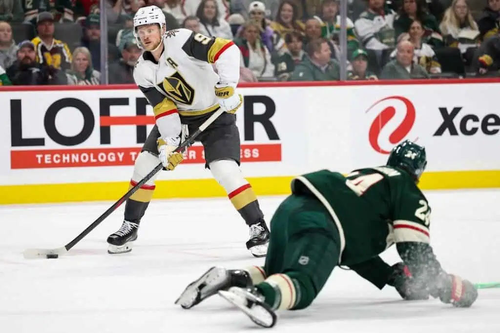 A Vegas Golden Knights player prepares to shoot the puck while a Minnesota Wild player sprawls on the ice, attempting to block the shot.