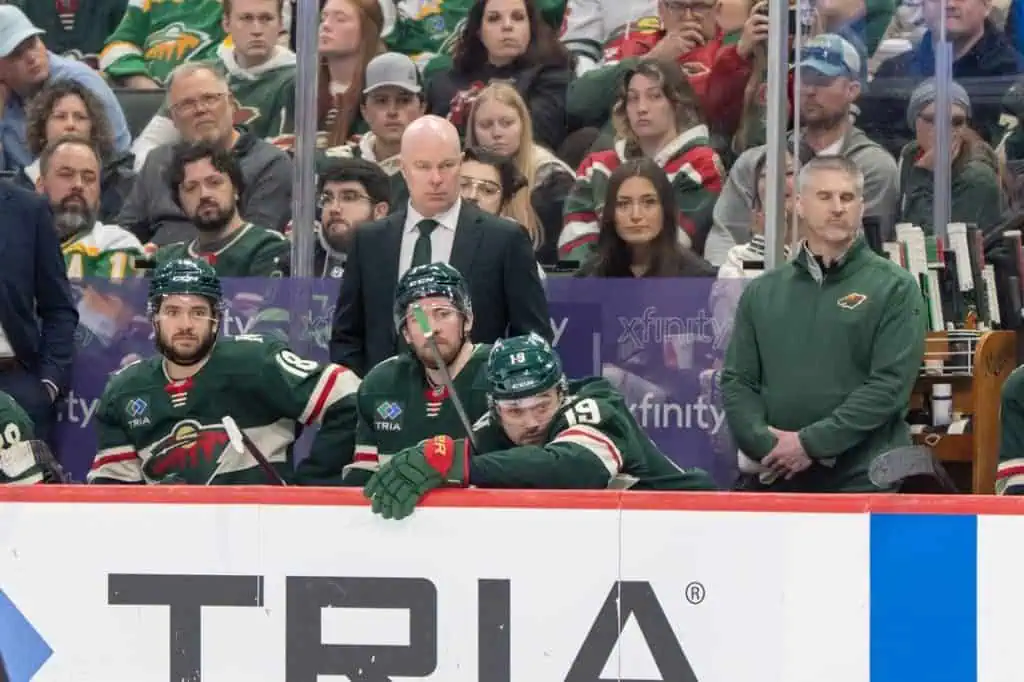 A group of hockey players in green jerseys sit on the bench, focused on the game, with a coach observing from the sidelines.
