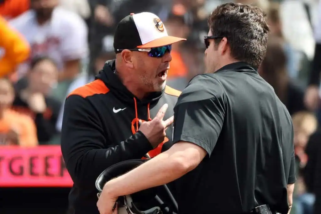 A coach in an orange and black hoodie gestures animatedly while conversing with an official at a baseball game.