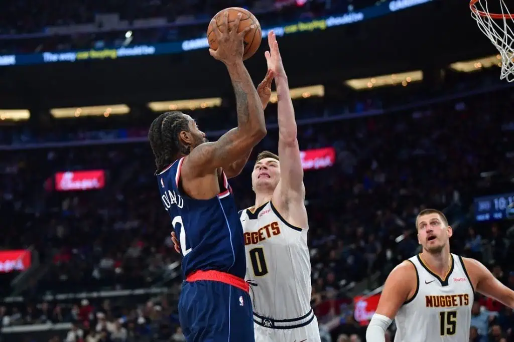 A basketball player in a navy uniform shoots over a defender in a white jersey during an intense game in an arena filled with spectators.