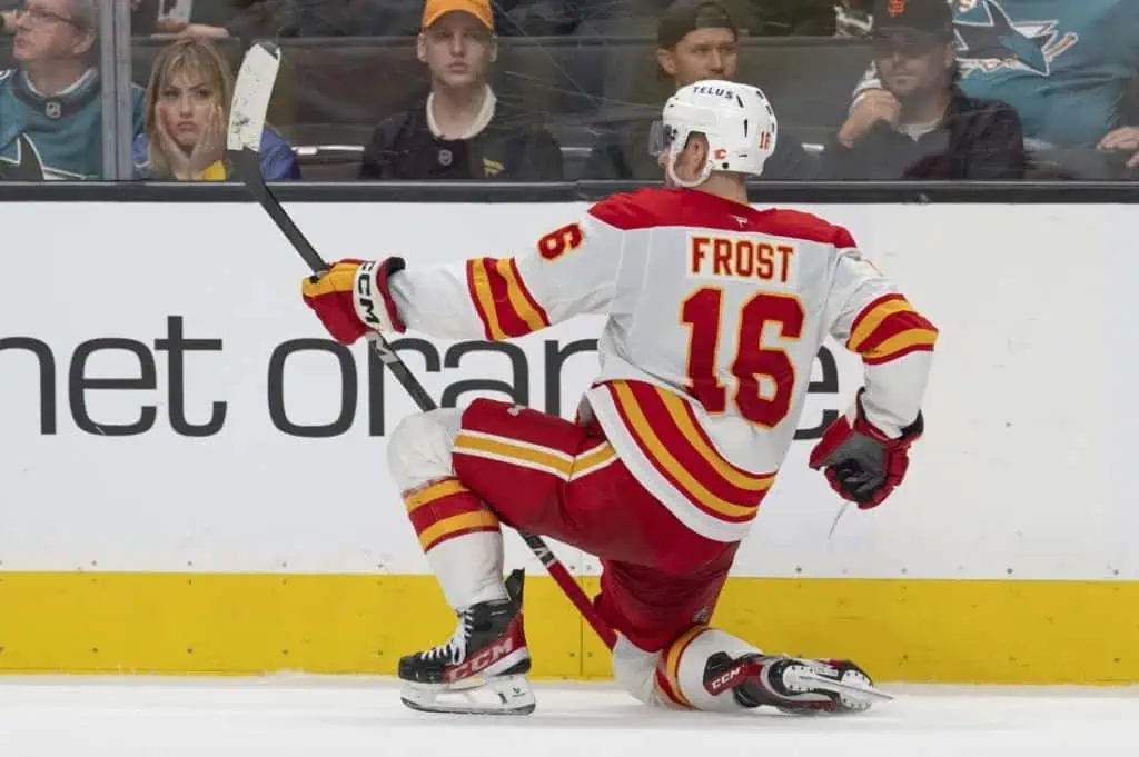 A hockey player in a red and white jersey with the name 'Frost' celebrates on the ice, one knee on the ground, stick raised triumphantly.