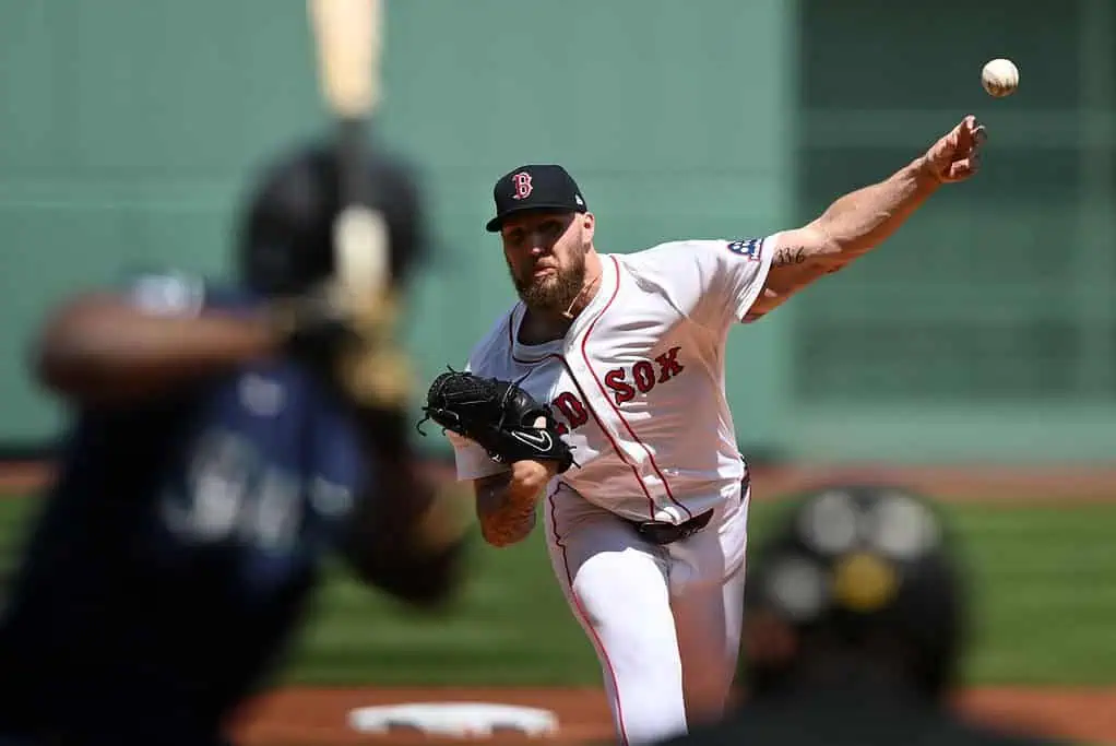 A pitcher in a red and white uniform throws a baseball, while a batter stands ready to swing in a sunny baseball stadium.