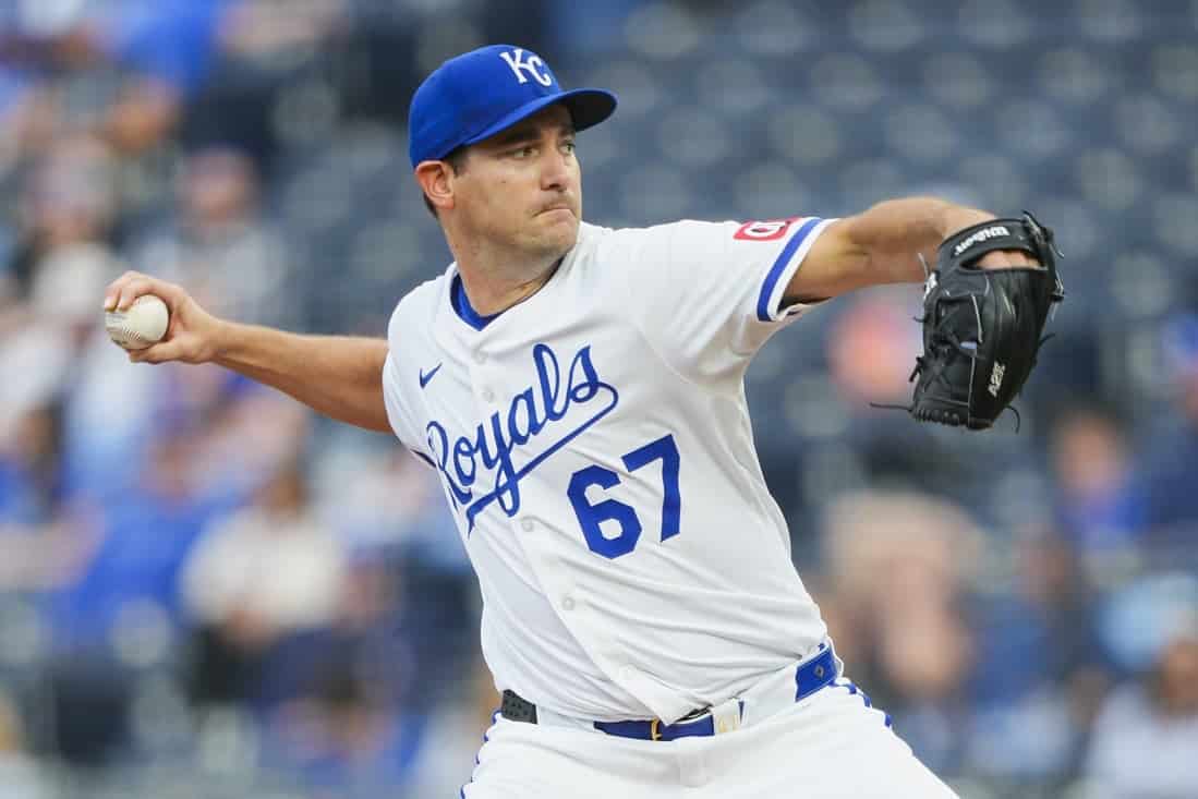 A Kansas City Royals player in a white jersey with blue lettering winds up to pitch a baseball against a blurred crowd in the background.