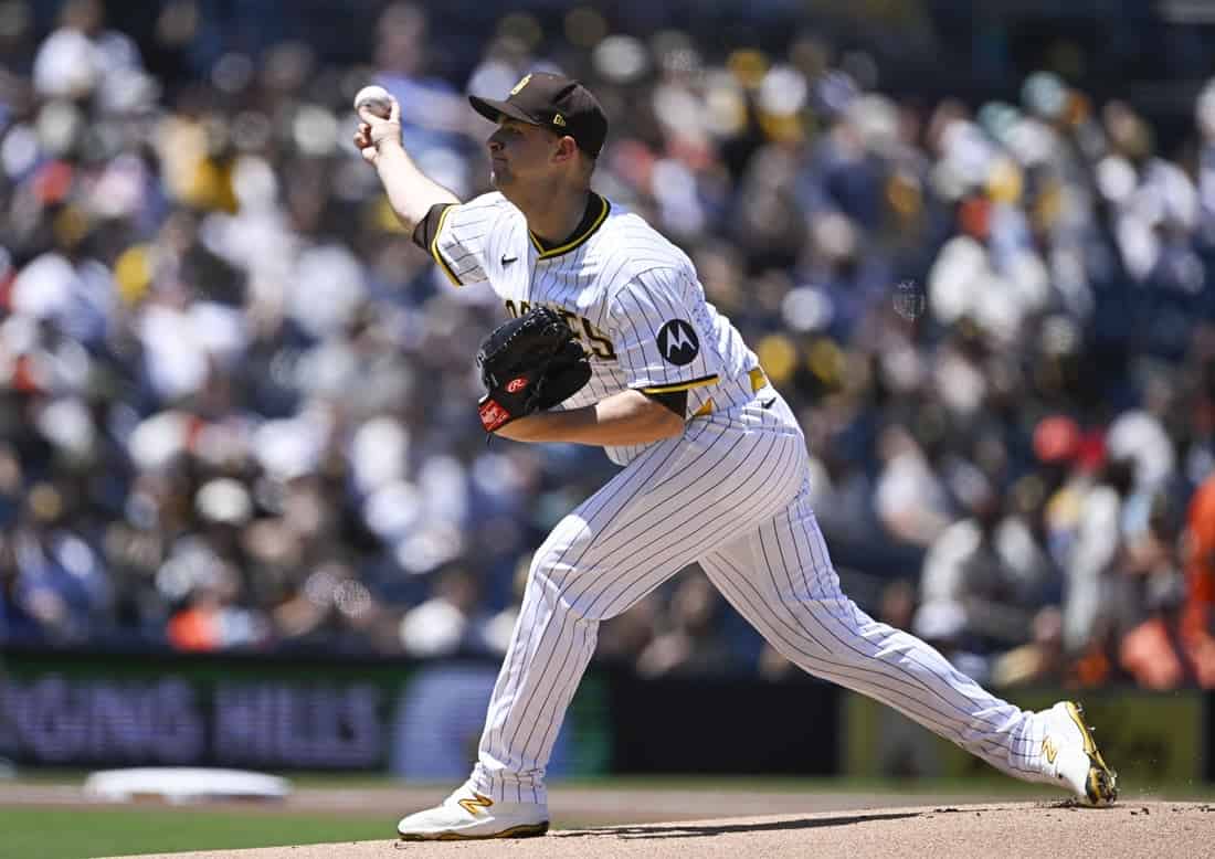 A baseball pitcher, wearing a white striped uniform, winds up to throw a ball during a sunny game, with a crowd in the background.