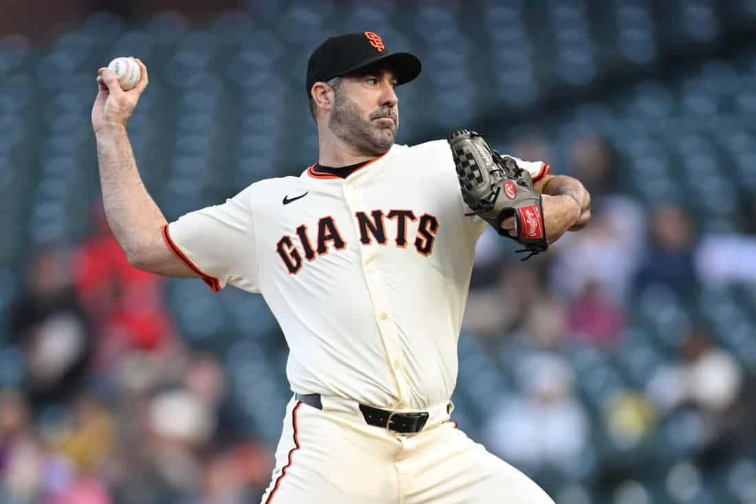 A pitcher in a San Francisco Giants uniform prepares to throw a baseball on a field, with blurred spectators in the background.