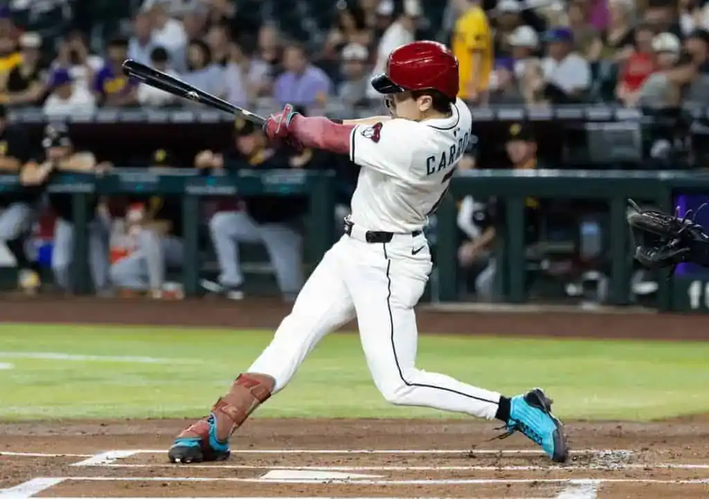 A baseball player swings a bat during a game, wearing a red helmet and a white uniform, with fans visible in the background.