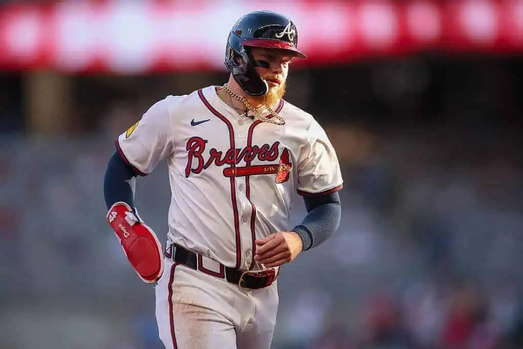 A baseball player in a white Braves jersey with a red stripe runs across the field, wearing a helmet and red batting gloves.