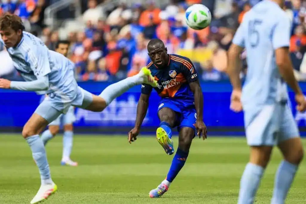 A soccer player in an orange and blue jersey strikes a flying ball, while an opposing player attempts a kick in a dynamic match scene.