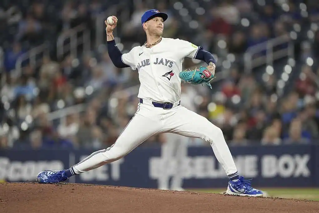 A baseball player in a Blue Jays jersey throws a pitch on the field during a night game, stadium lights illuminating the action.