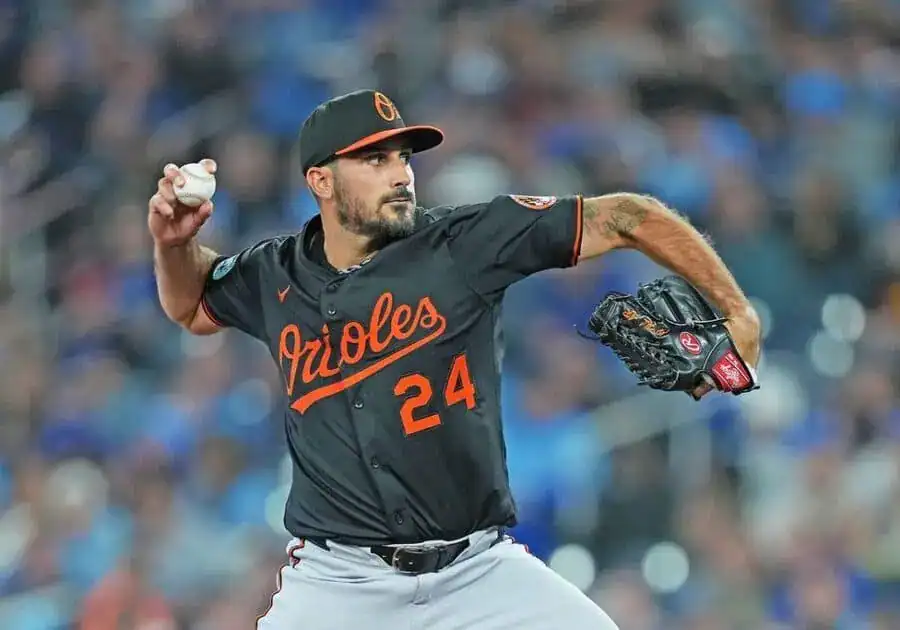A Baltimore Orioles pitcher winds up to throw a baseball, wearing a black jersey with orange accents and a cap, focused on the game.