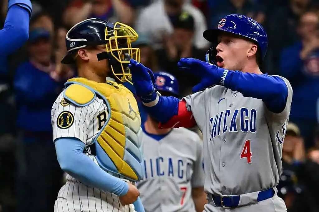 A Chicago Cubs player celebrates after scoring, while a catcher in a striped uniform watches, amidst a lively crowd.