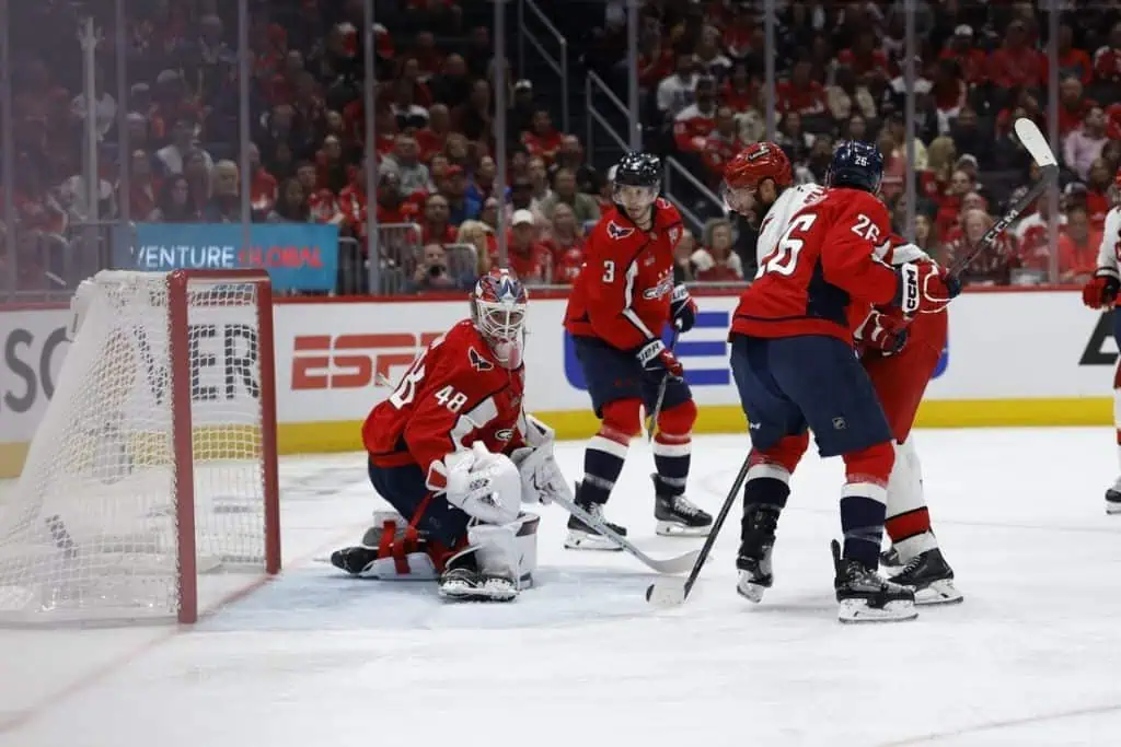 A tense moment in a hockey game, with players in red jerseys battling near the goal while the goalie is ready in the net.