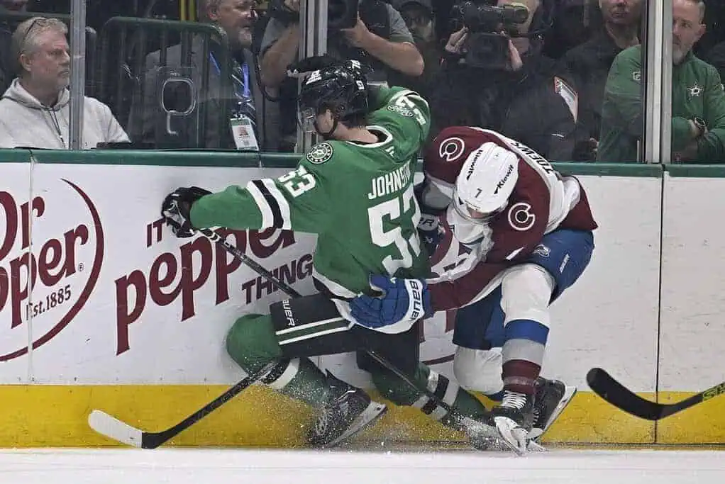A hockey player in a green jersey is pinned against the boards by an opponent in a maroon and white jersey during a game.