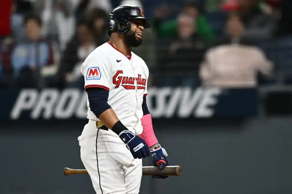 7 A baseball player wearing a Guardians jersey stands on the field, holding a bat and wearing pink batting gloves, ready to play.