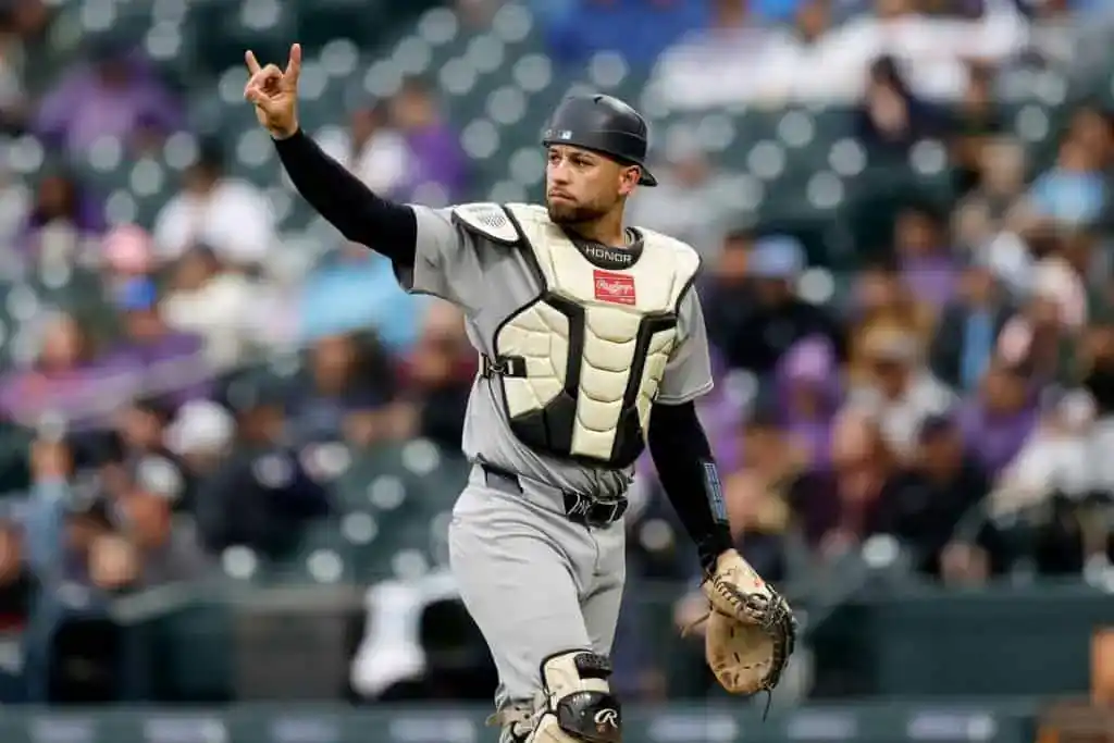 A baseball catcher in a gray uniform signals to players from the pitcher's mound during a game, with a stadium crowd visible in the background.