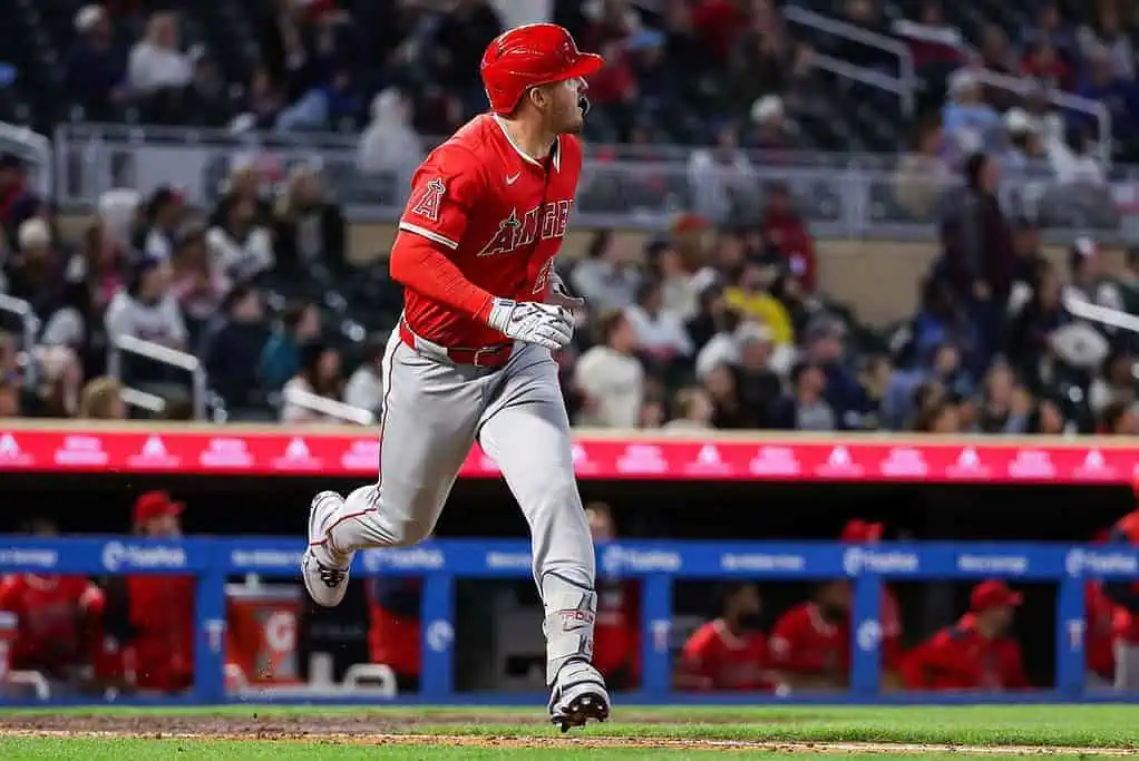 A baseball player in a red uniform is running towards first base, with a blurred crowd of fans