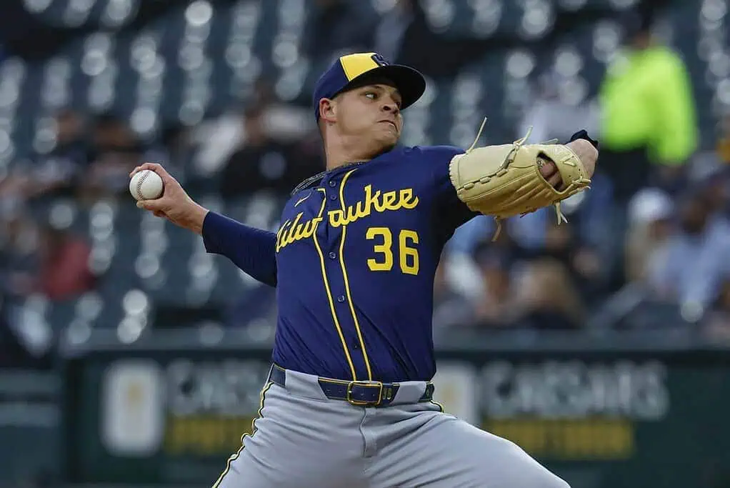 A baseball pitcher in a blue and yellow uniform throws a ball, with a focused expression, on a field filled with spectators.