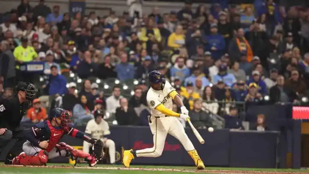 A baseball player in a yellow and white uniform swings at a pitch, with fans and stadium lights visible in the background.
