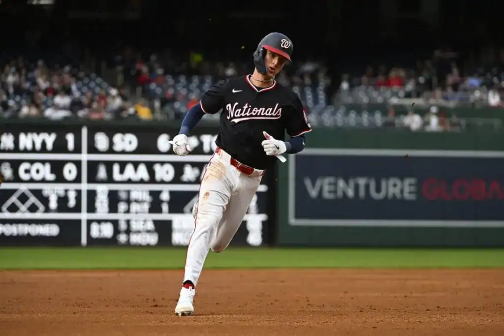 A player in a black Nationals jersey runs on the baseball field, with a scoreboard displaying game scores in the background.