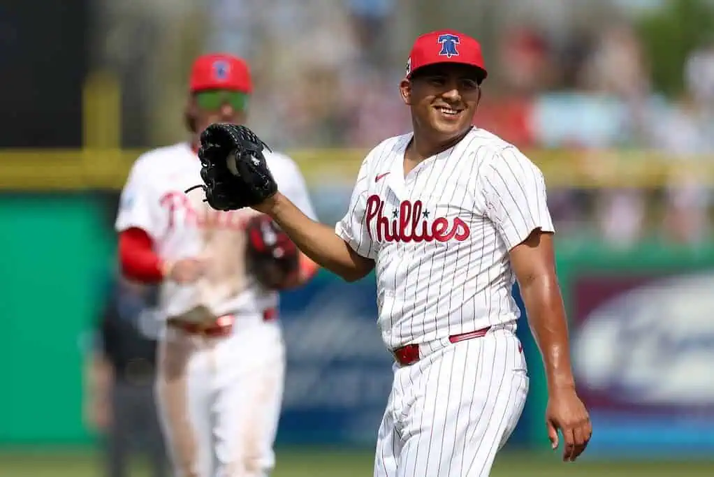 A Philadelphia Phillies player in a striped jersey and red cap walks on the field holding a baseball glove, with cheering fans in the background.