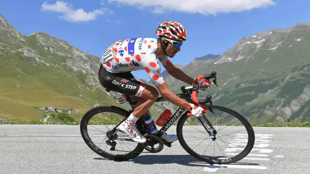 A professional cyclist in a polka-dot jersey rides a sleek black bicycle on a winding mountain road with green hills and blue sky.
