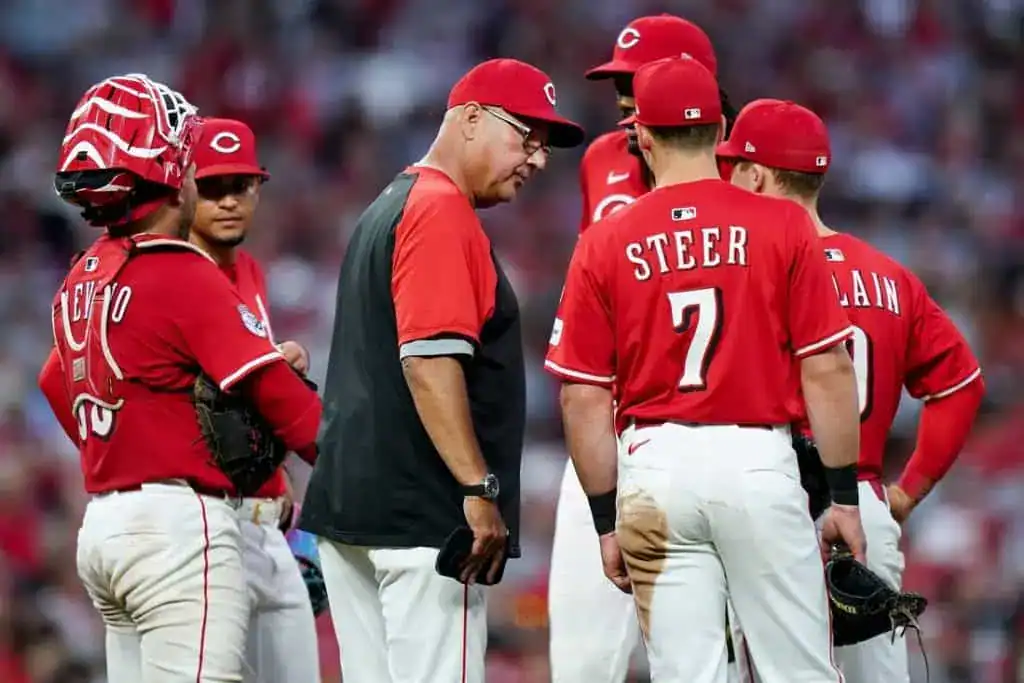 A group of Cincinnati Reds players and a coach gather on the field, discussing strategy during a baseball game.