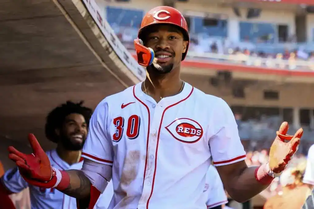 A baseball player in a Reds jersey stands in the dugout with a confident expression, gesturing with his hands as teammates observe.