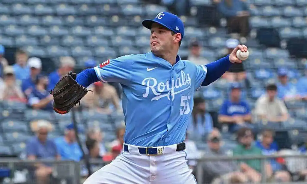A pitcher in a light blue Kansas City Royals jersey throws a baseball on a sunny day, with spectators blurred in the background.