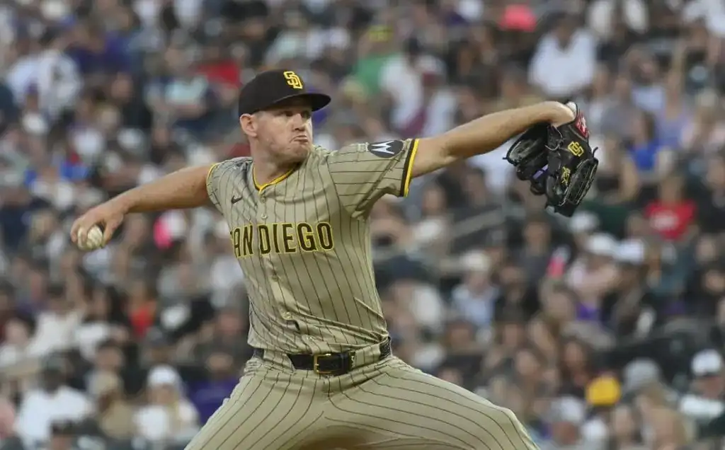 A pitcher in a tan San Diego Padres uniform winds up to throw a baseball, with a blurred crowd of fans cheering in the background.