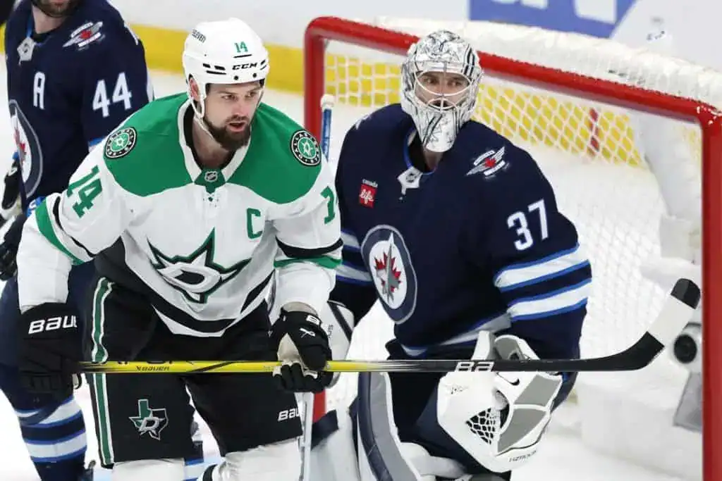 A hockey player in a green and white jersey stands in front of the net, while another player in a dark blue uniform prepares to defend.