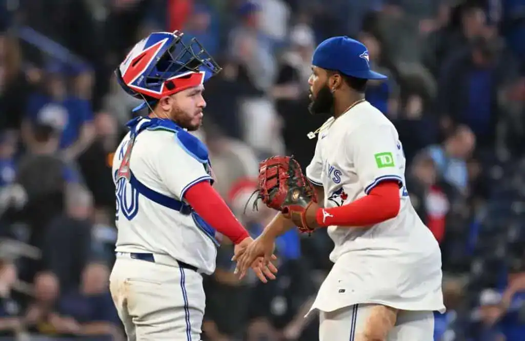 Two baseball players in Toronto Blue Jays uniforms celebrate on the field, surrounded by cheering fans in the stadium.