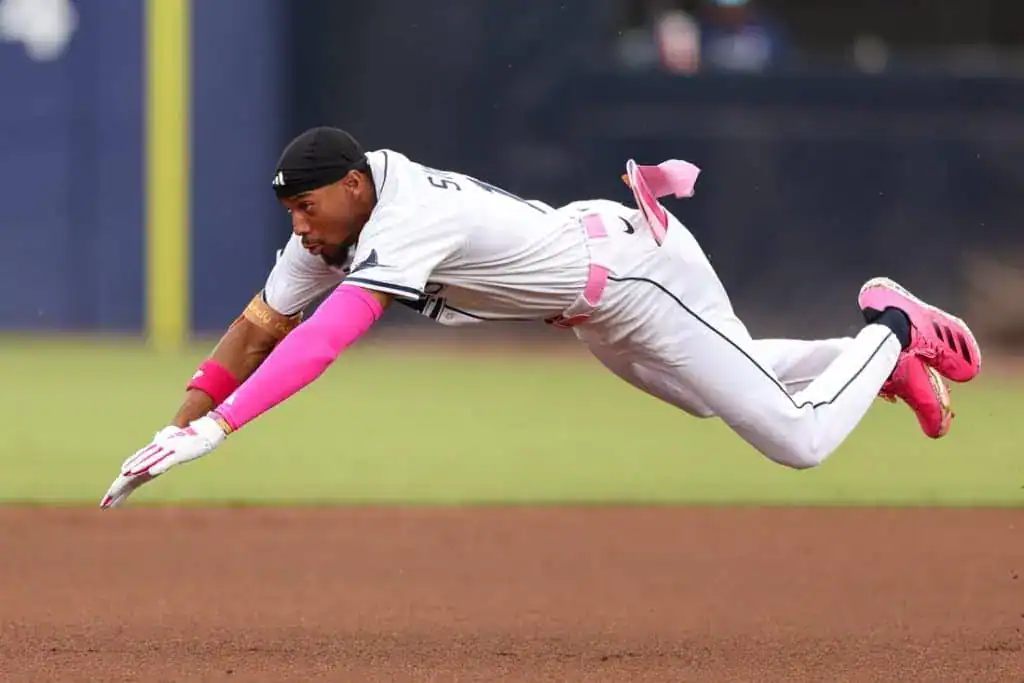 A baseball player dives for a ground ball, wearing a white uniform with pink accents and bright pink cleats on a grassy field.
