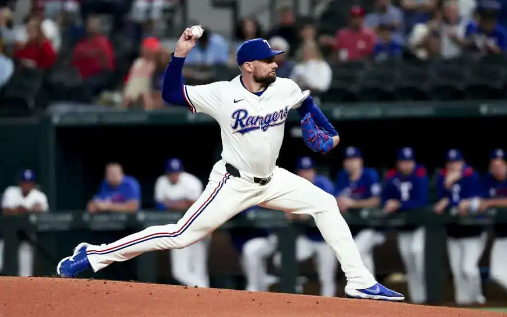A pitcher in a Texas Rangers uniform winds up for a throw on the mound, surrounded by teammates in blue jerseys in the background.